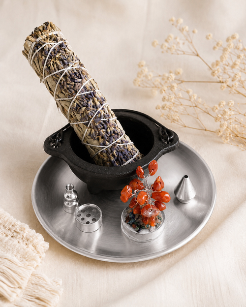 Silver energy-clearing tray with a black cauldron, dried herb bundle, orange gemstone tree, and incense-holder pieces.