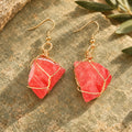 Pair of coral-red rough stone earrings with gold-tone wire wrapping on a warm stone surface.