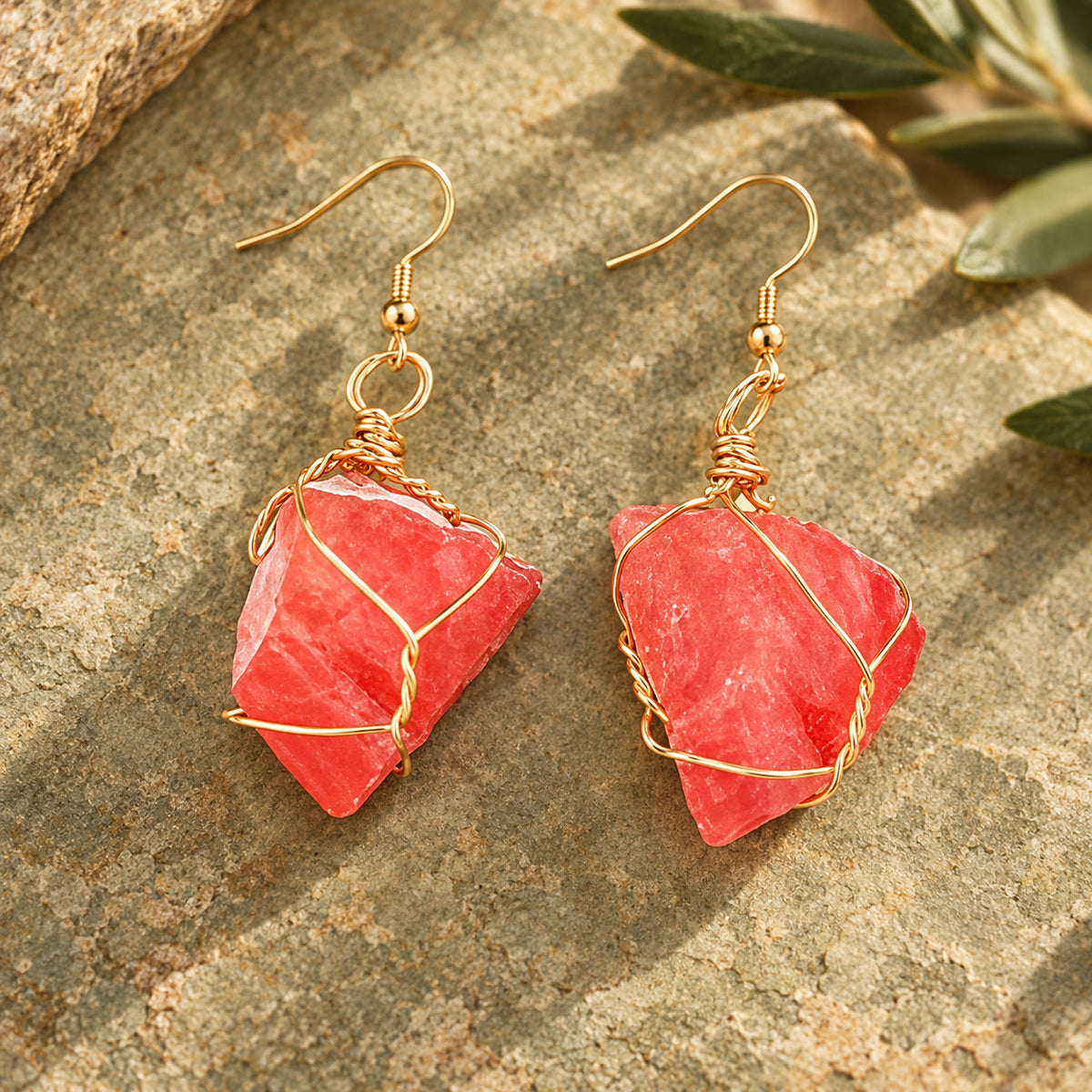 Pair of coral-red rough stone earrings with gold-tone wire wrapping on a warm stone surface.