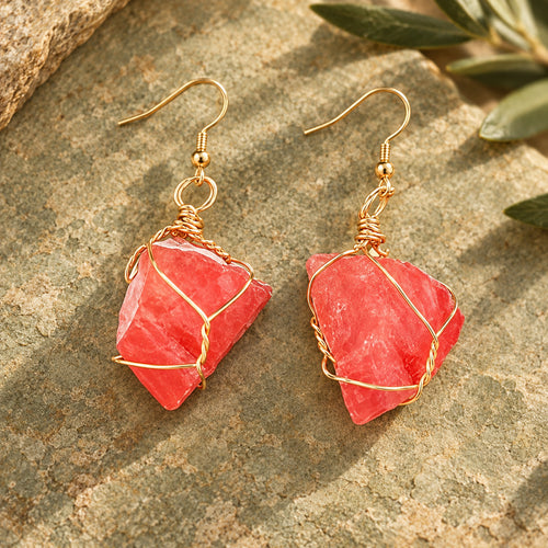 Pair of coral-red rough stone earrings with gold-tone wire wrapping on a warm stone surface.