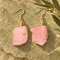 Pair of soft pink rough stone earrings with gold-tone wire wrapping on a warm stone surface.