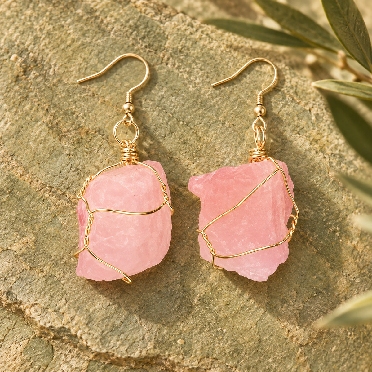 Pair of soft pink rough stone earrings with gold-tone wire wrapping on a warm stone surface.