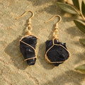 Pair of glossy black rough stone earrings with gold-tone wire wrapping on a warm stone surface.