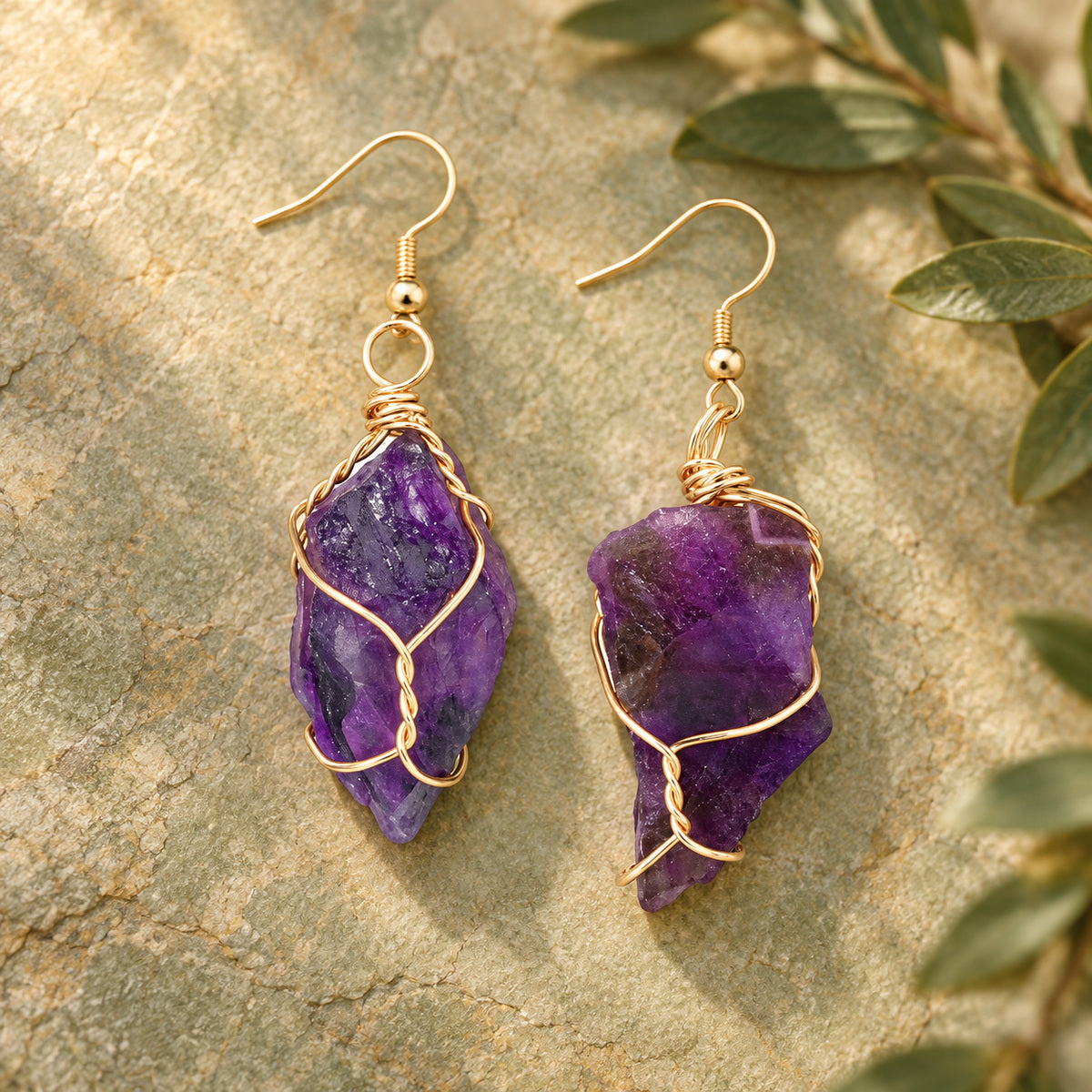 Pair of deep purple rough stone earrings with gold-tone wire wrapping on a warm stone surface.