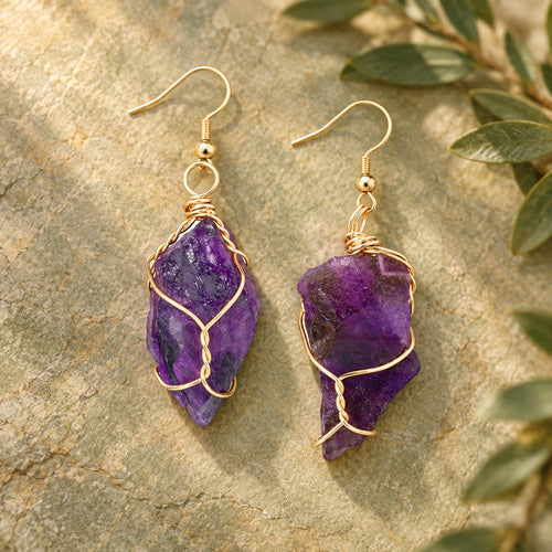 Pair of deep purple rough stone earrings with gold-tone wire wrapping on a warm stone surface.